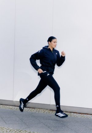 Chaqueta negra con cremallera y capucha, leggins negros y zapatillas deportivas negras con franjas blancas. Las zapatillas y la ropa tienen un diseño deportivo.