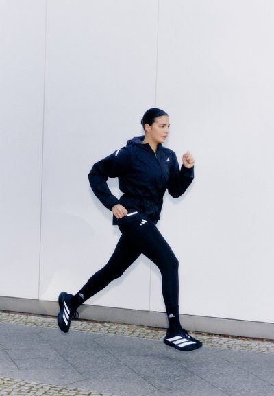 Chaqueta negra con cremallera y capucha, leggins negros y zapatillas deportivas negras con franjas blancas. Las zapatillas y la ropa tienen un diseño deportivo.