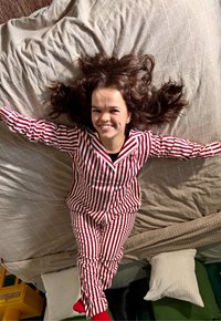 Femme souriante aux longs cheveux bouclés allongée sur le lit, les bras étendus, portant un pyjama rayé rouge et blanc ainsi que des chaussettes rouges.