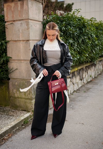 Mujer de pie al aire libre, vestida con chaqueta de cuero negra, blusa blanca, corsé gris, pantalones anchos negros, zapatos burdeos y sosteniendo un bolso burdeos.
