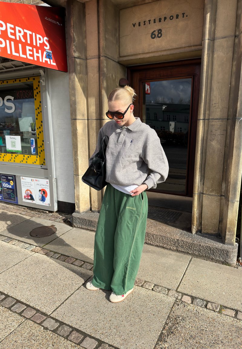 Personne avec des lunettes de soleil, un tee-shirt gris à manches longues, un pantalon large vert et un sac noir, debout près d'une porte en pierre portant l'inscription « Vesterport 68 » en plein jour.