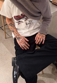 Άτομο καθισμένο που φοράει μαύρο παντελόνι, γυαλιστερά μαύρα παπούτσια, λευκό T-shirt με γραφικό σχέδιο, γκρι πλεκτή ζακέτα που κρέμεται από τον ώμο, με τατουάζ στα χέρια και ασημένια δαχτυλίδια.