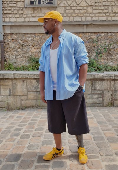 Camisa a rayas azul y blanca sobre una camiseta blanca, pantalones cortos negros y zapatillas amarillas con detalles en negro. Presenta una pierna prostética metálica.