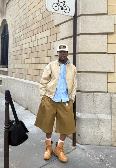 Chaqueta ligera beige sobre una camisa azul claro, combinada con pantalones cortos marrones y botas canela. Una bolsa negra descansa apoyada en un poste cercano.