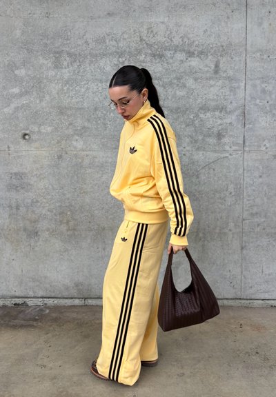 Mujer con chándal amarillo de Adidas con franjas negras sostiene un bolso marrón tejido, de pie contra una pared de hormigón gris, mirando hacia abajo.