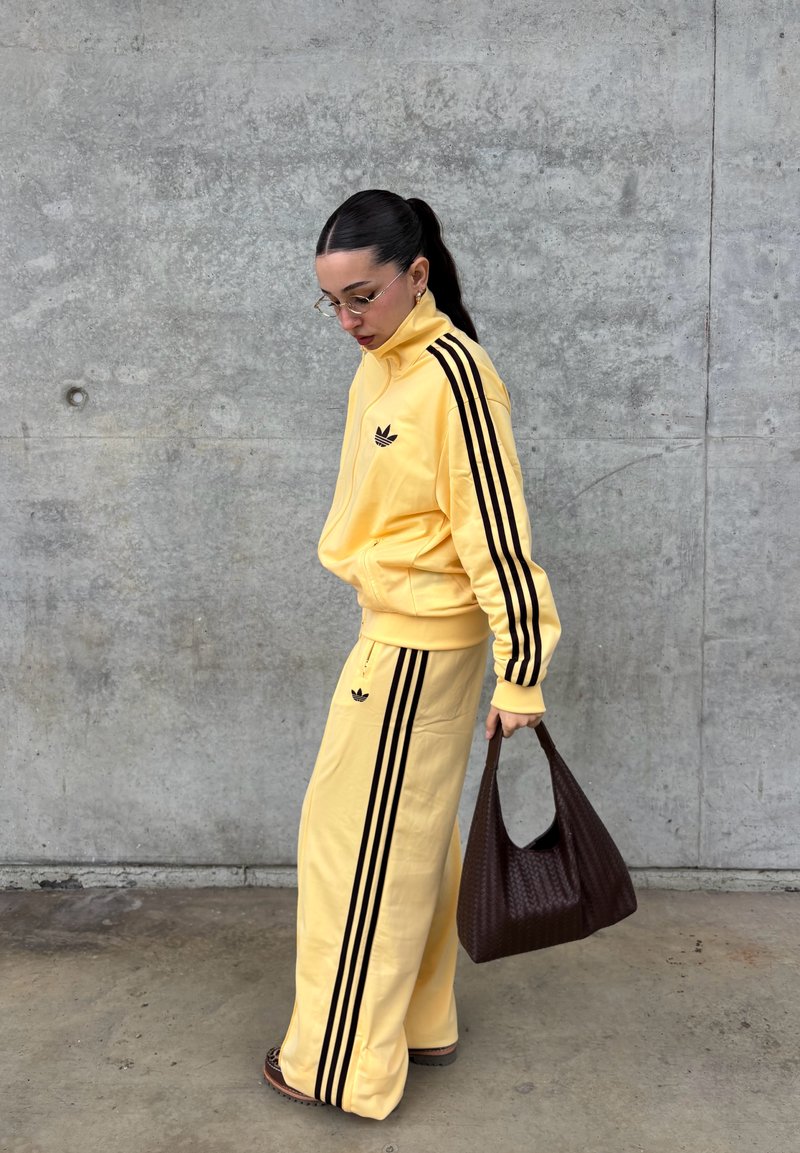 Femme en survêtement Adidas jaune à rayures noires tenant un sac à main en osier marron, debout contre un mur en béton gris, regardant vers le bas.
