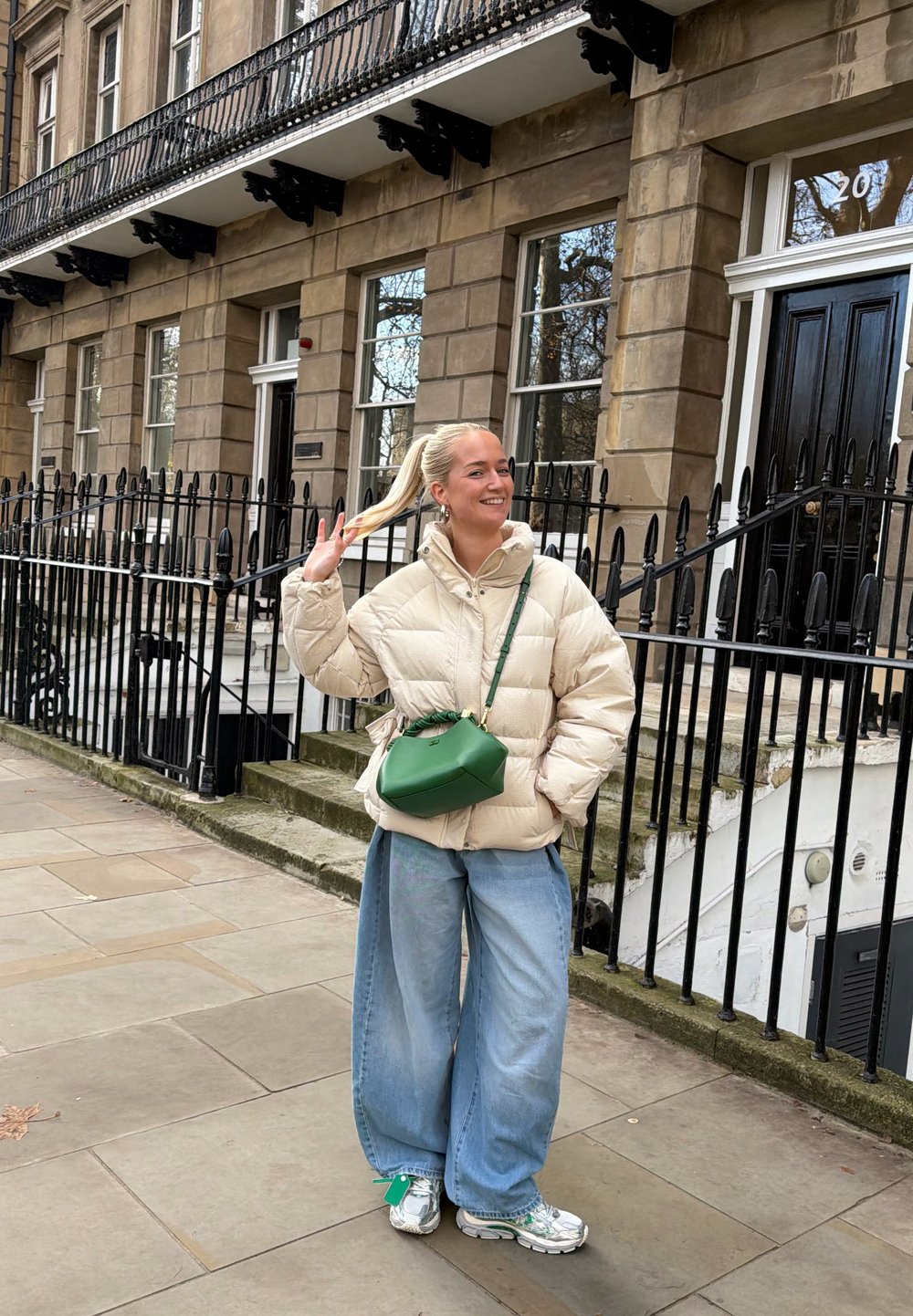Veste matelassée beige, large jean bleu clair, et sac à main vert. Des baskets élégantes avec des accents argentés complètent la tenue. Décor urbain avec des rambardes.