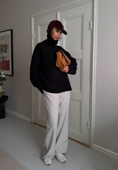 Mujer con suéter de cuello alto negro, pantalones anchos beige, gorra burdeos, zapatillas blancas, sosteniendo un bolso marrón claro, de pie en el interior cerca de una puerta blanca cerrada.