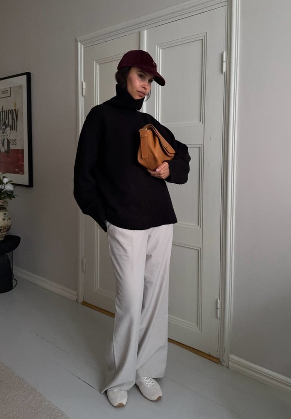 Femme portant un col roulé noir, un pantalon large beige, une casquette bordeaux, des baskets blanches, tenant un sac à main beige, debout à l'intérieur près d'une porte blanche fermée.