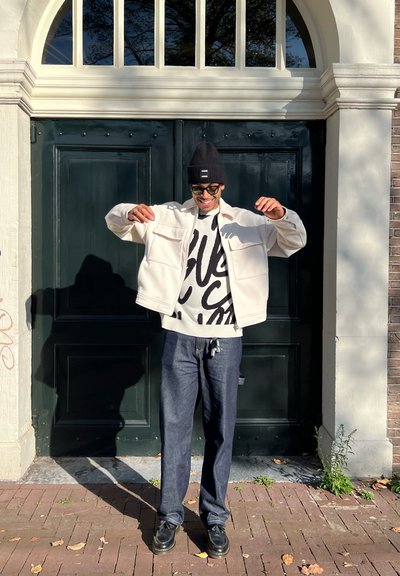 Chaqueta blanca con dos bolsillos frontales, suéter gráfico negro, jeans azul oscuro, zapatos negros y un gorro negro, de pie frente a puertas verdes.