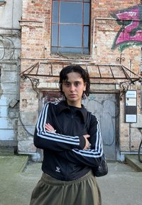 Fekete cipzáras dzseki fehér csíkokkal, hosszú olívazöld szoknyával párosítva. Ezüst karika fülbevalók és egy kis fekete táska. Téglafal háttér.