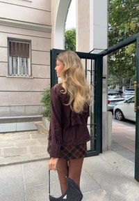 Braune Bluse mit einem Rückensaum, kombiniert mit einem karierten Rock. Als Accessoire dient eine schwarze strukturierte Handtasche. Set im Freien in der Nähe eines Torbogens.