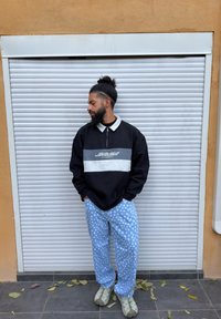 Homme avec barbe et chignon portant un sweat-shirt noir et gris, un pantalon bleu clair à pois et des baskets, debout les mains dans les poches contre un rideau métallique.