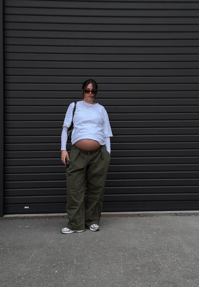 Mujer embarazada con gafas de sol, camiseta blanca de capas y pantalones verdes holgados, de pie frente a una puerta de garaje de metal oscuro, sosteniendo un teléfono en la mano derecha.