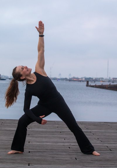 Mujer vestida con ropa deportiva negra realizando la postura de yoga de ángulo lateral extendido en un muelle de madera junto al agua bajo un cielo nublado.