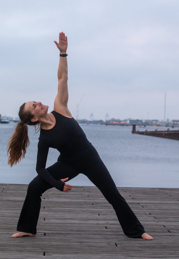Kvinne i svart treningstøy utfører utvidet sidevinkel-yogaposisjon på en trebru ved vannet under overskyet himmel.