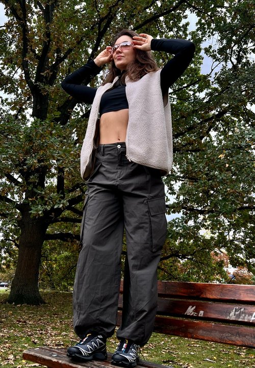 Pantalons cargo gris avec plusieurs poches, un gilet en polaire beige sur un crop top noir, et des chaussures de randonnée noires, disposés sur un banc en bois à l'extérieur.