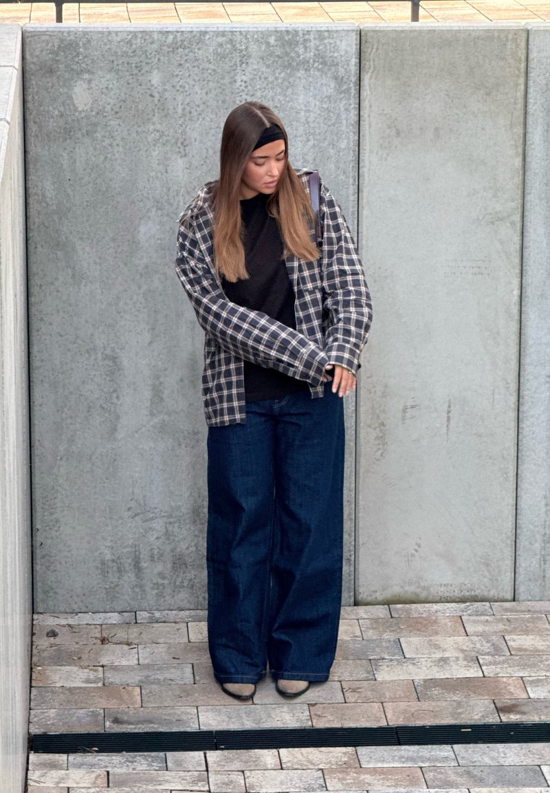 Pledēta flaneļa krekls melnā un pelēkā krāsā, valkāts pār melnu topu; kombinēts ar platām tumši zilām džinsu biksēm. Teksturētas virsmas, minimāls aksesuāru daudzums.