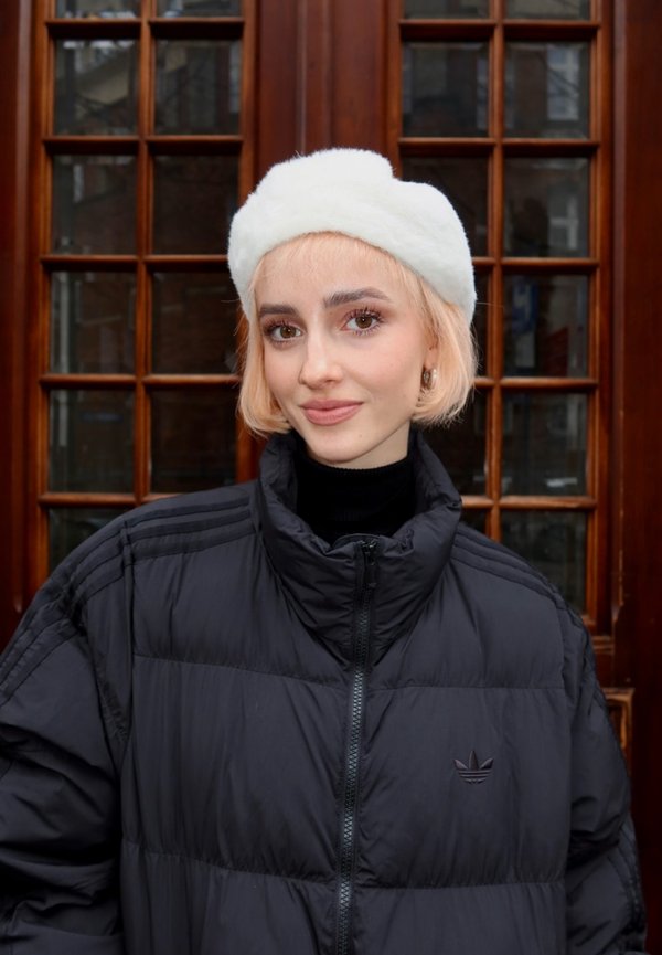 Ung kvinde med kort blondt hår iført hvidt, fluffy hat og sort dynejakke, stående foran en trædør med vinduer.