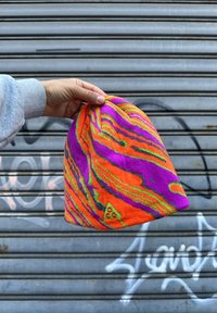 Main tenant un bonnet tricoté coloré avec des tourbillons orange, violet, vert et noir devant un volet métallique gris recouvert de graffiti.