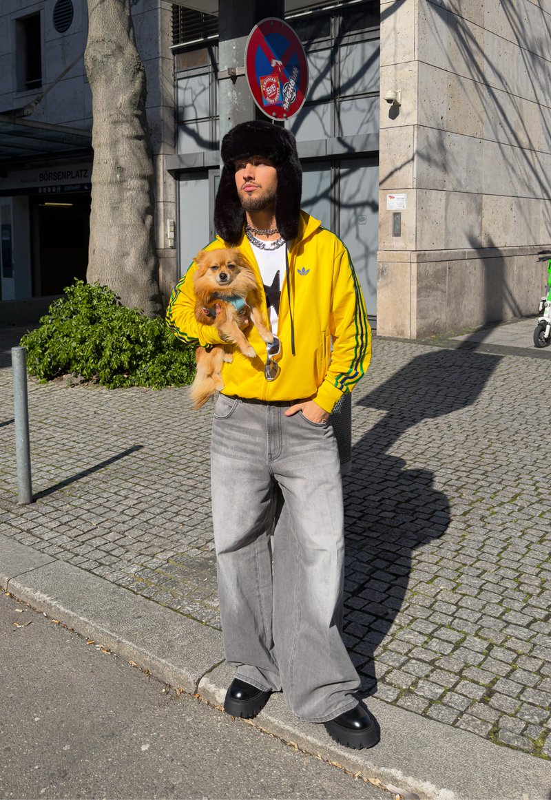 Man in yellow jacket and black fur hat holding small dog on city sidewalk near no-parking sign on sunny day.
