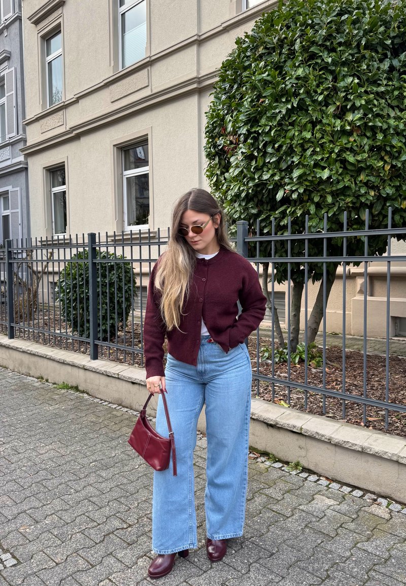 Jeune femme portant un cardigan bordeaux, un jean large bleu, des bottes bordeaux assorties et un sac à main, debout sur un trottoir en ville.