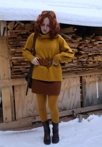 Femme aux cheveux roux portant un pull moutarde, une jupe marron et des bottes, ajustant une large ceinture, debout sur la neige devant un tas de bûches empilées.