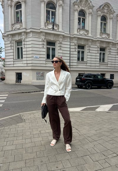 Blazer de lino blanco de doble botonadura, pantalones anchos marrones, sandalias claras y un bolso negro, con un fondo de calle urbana.