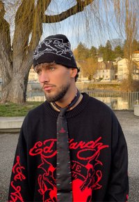 Homme barbu portant un bonnet noir à motifs, un pull noir avec un texte graphique rouge, et une cravate noire, debout à l'extérieur près d'un arbre et d'un plan d'eau.