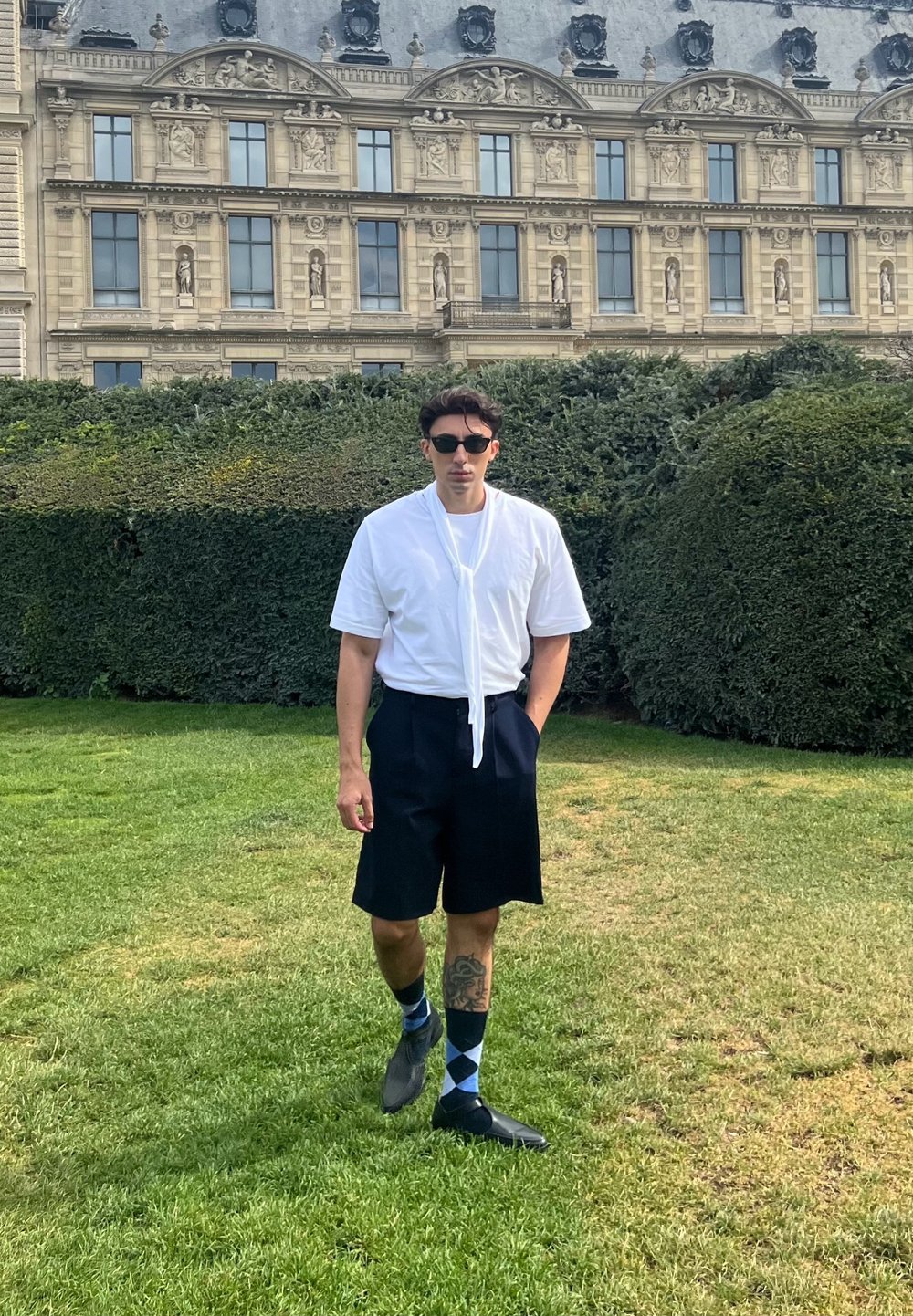 Camicia bianca a maniche corte con cravatta, pantaloni corti scuri, scarpe nere e calzini blu a rombi; in piedi in un'area erbosa con siepi.
