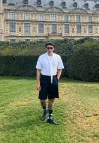 Camicia bianca a maniche corte con cravatta, pantaloni corti scuri, scarpe nere e calzini blu a rombi; in piedi in un'area erbosa con siepi.