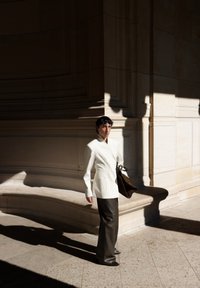 Hvit skreddersydd blazer med dyp V-utringning, kombinert med mørkegrønne vide bukser. Modellen holder en brun skulderveske mot en steinbakgrunn.