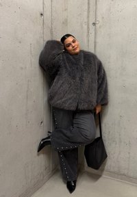 Veste en fausse fourrure grise à manches larges, associée à un pantalon en denim foncé orné de clous métalliques. Bottines noires à bout pointu et sac à main noir à épaule.