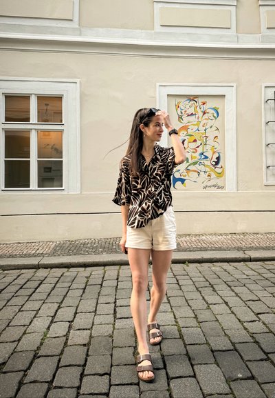 Camisa de corte holgado en patrones negros y beige, pantalones cortos blancos y sandalias marrones. Caminando por una calle de adoquines cerca de un edificio de color claro y un mural.