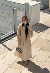 Bežinė bombakinė striukė su juodu apykakle, permatomas juodas marškinėlis ir prigludusi bežinė sijonas. Priedai apima raštuotą rankinę ir saulės akinius.