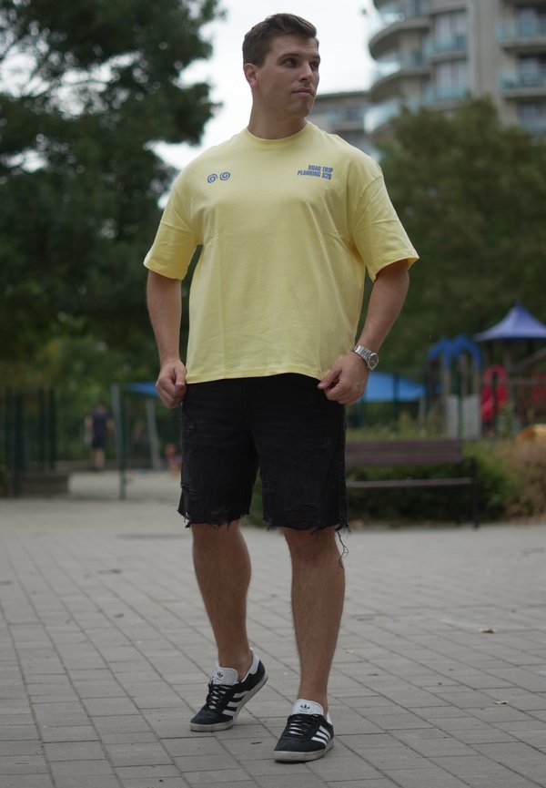 Ein Mann in einem gelben T-Shirt und schwarzen Shorts steht auf einem gepflasterten Weg in einem Park mit Bäumen und Spielplatzgeräten im Hintergrund.
