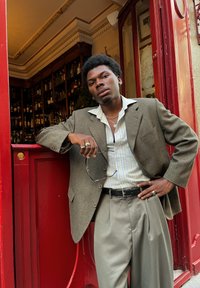 Blazer en laine gris porté sur une chemise à rayures claires, avec un pantalon plissé gris clair. Les accessoires comprennent des bagues et des lunettes, le tout sur fond rouge.
