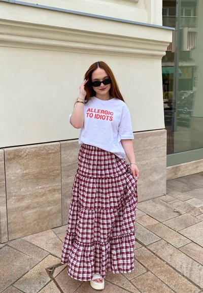 Camiseta blanca con texto rojo que dice "ALÉRGICA A LOS IDIOTAS" combinada con una falda larga de cuadros rojos y blancos con múltiples capas. Sandalias blancas completan el atuendo.