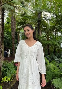 Abito bianco di lunghezza al ginocchio con dettagli floreali a intaglio, maniche a palloncino e chiusura frontale con bottoni, ambientato su uno sfondo tropicale verde lussureggiante.