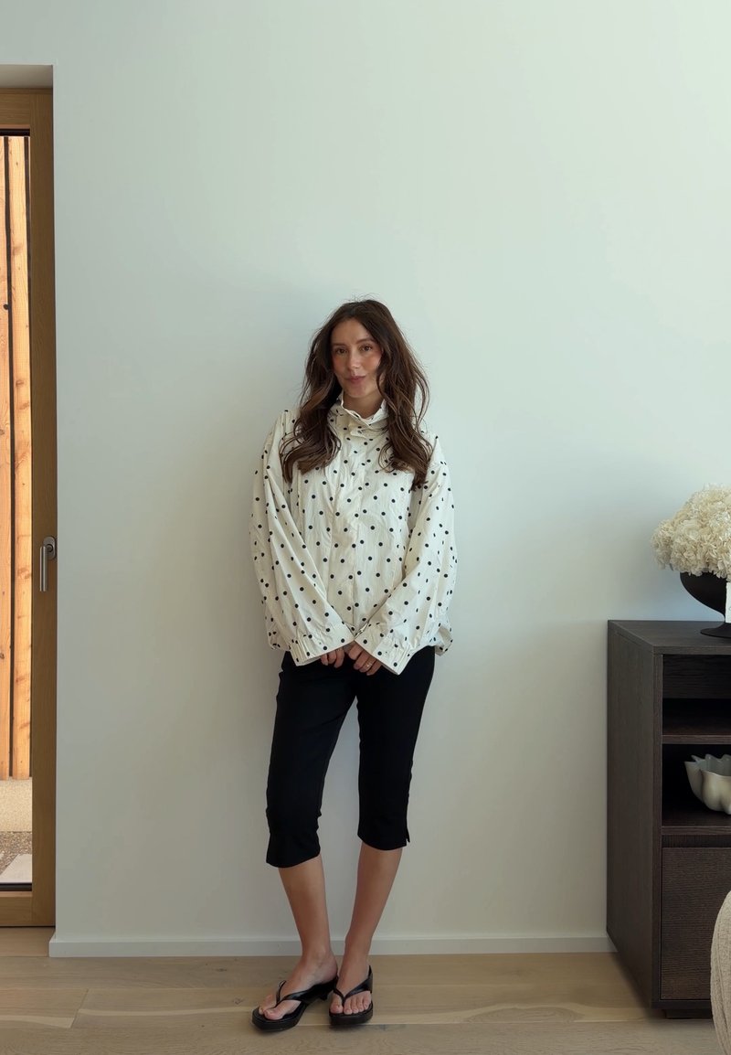 Femme aux longs cheveux foncés portant un chemisier blanc à pois noirs et un pantalon capri noir, debout pieds nus dans une pièce minimaliste.