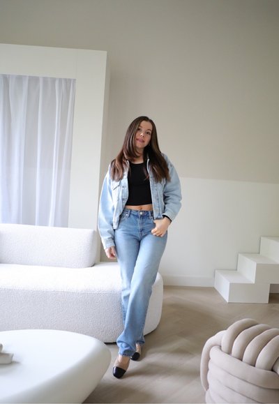 Chaqueta de mezclilla azul claro sobre un top corto negro, combinada con jeans azul claro. El entorno cuenta con un sofá blanco texturizado y un suelo de madera clara.