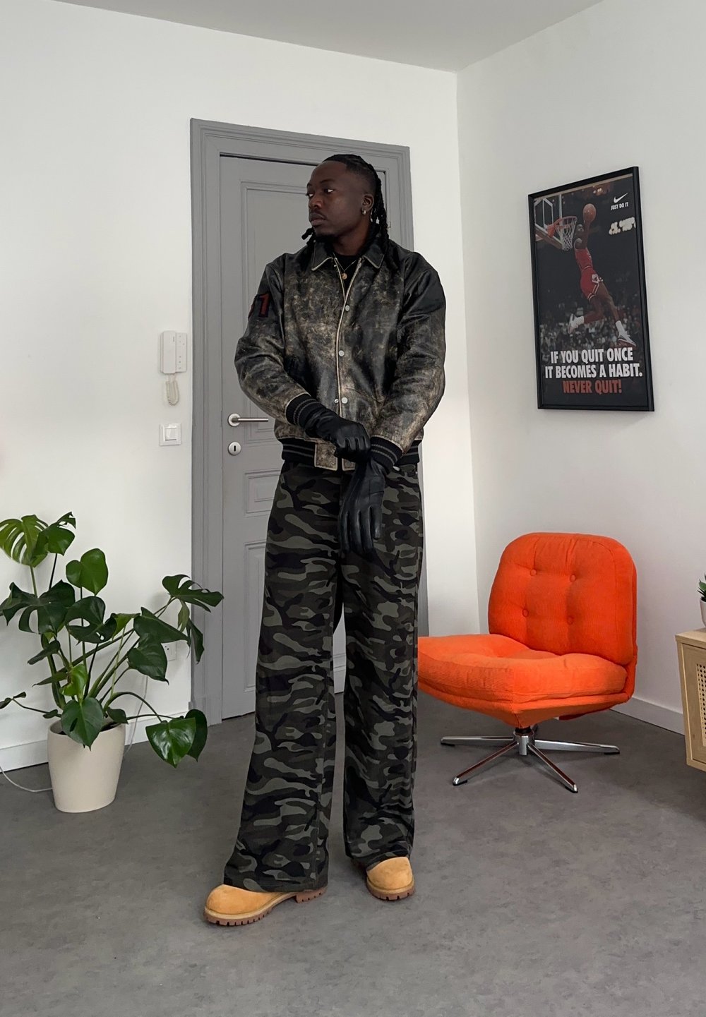 Homme portant une veste en cuir noire, un pantalon camouflage, des bottes beige et des gants noirs, debout dans une pièce avec une chaise orange et une affiche motivante de basketball.