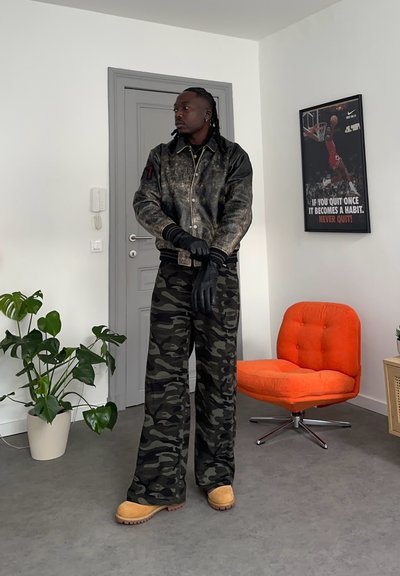 Hombre con chaqueta de cuero negra, pantalones de camuflaje, botas color beige y guantes negros de pie en una habitación con una silla naranja y un cartel motivacional de baloncesto.