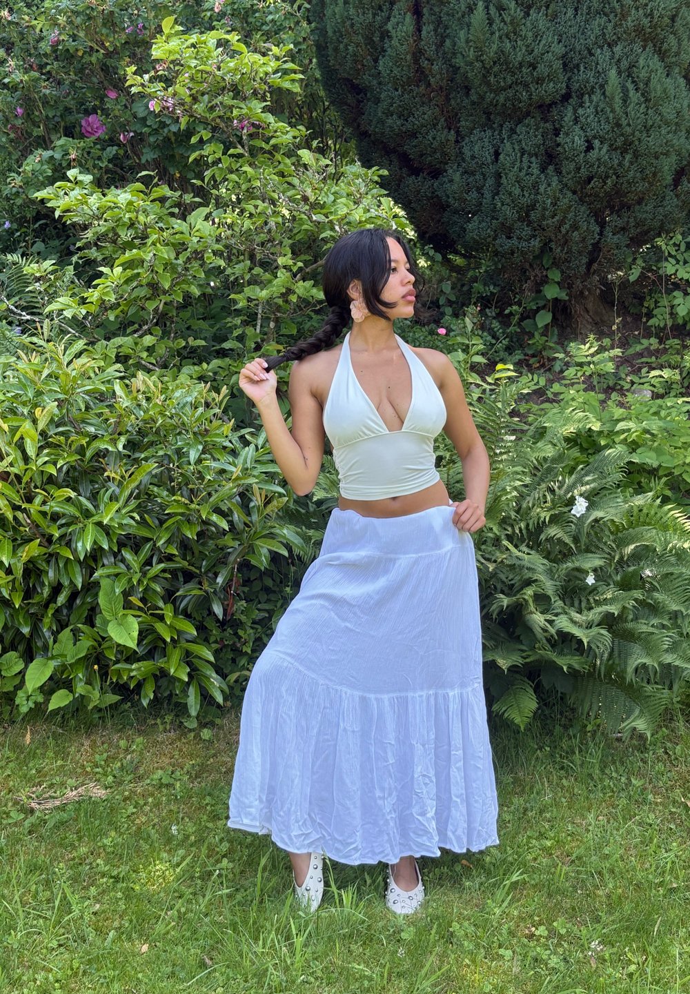 Biały top na halter i długa biała spódnica z falbanką. W tle zielone liście i rośliny. Jasne naturalne światło.