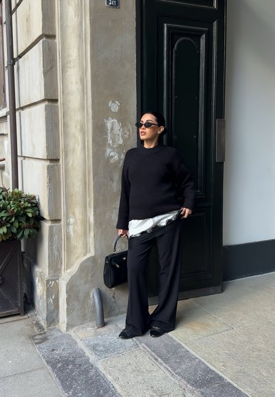 Mujer con suéter y pantalones negros, sosteniendo un bolso negro, de pie junto a una pared beige desgastada y una puerta de madera oscura en una calle empedrada.