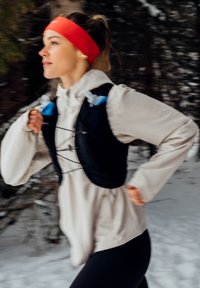 Femme courant en plein air dans une forêt enneigée, portant une veste beige, un gilet d'hydratation noir et un bandeau orange vif.