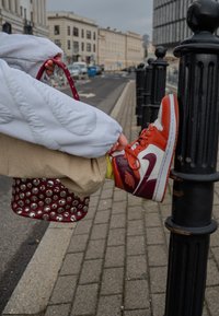 Pessoa ajusta um ténis alto vermelho e branco enquanto está sentado na calçada da cidade, segurando uma bolsa marrom com studds.