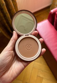 Bronzer compatto in una custodia rotonda con specchio riflettente, con una polvere marrone chiaro liscia e un'incisione del logo.