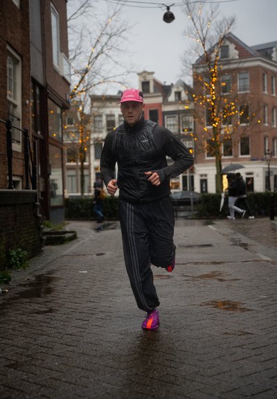 Hombre corriendo en una calle de adoquines mojados, vestido con ropa deportiva oscura y un sombrero rosa, con árboles envueltos en luces de cuerda naranjas y edificios al fondo.