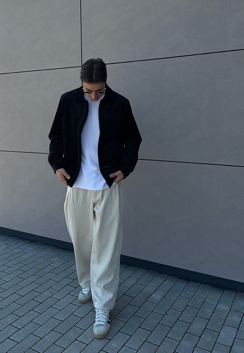 Homme portant des lunettes de soleil, une veste noire, une chemise blanche et un pantalon beige, debout sur un trottoir gris devant un mur lisse gris, regardant vers le bas.